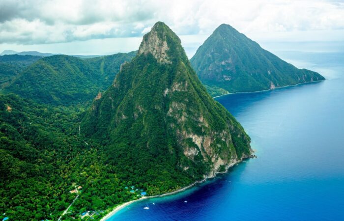 The-Pitons-Saint Lucia Aerial view of lush green mountains along a turquoise coastline with small houses at the shore and boats on calm blue water, tropical scenery.